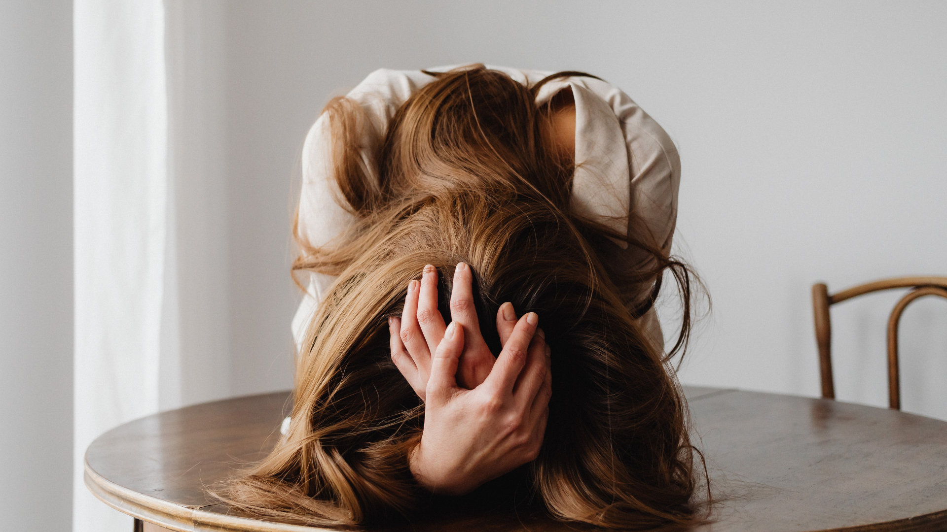 Blog 7 A woman sitting at a wooden table, bent forward with her head down and hands in her hair, appearing overwhelmed by financial stress.